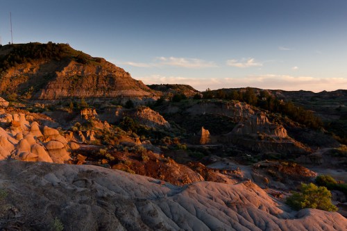 Sunset over rugged hills and rock formations, casting warm light on the landscape with scattered vegetation.