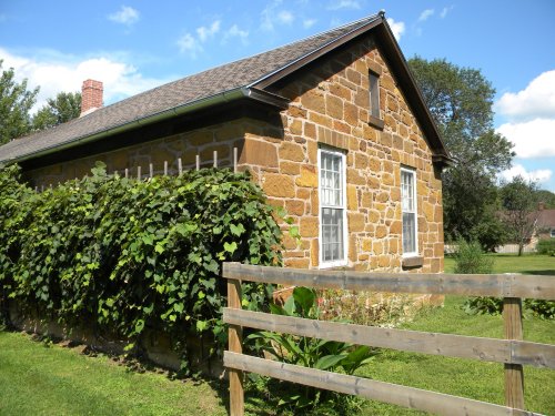 A stone house with a sloped roof, surrounded by greenery and a wooden fence, under a blue sky with clouds.