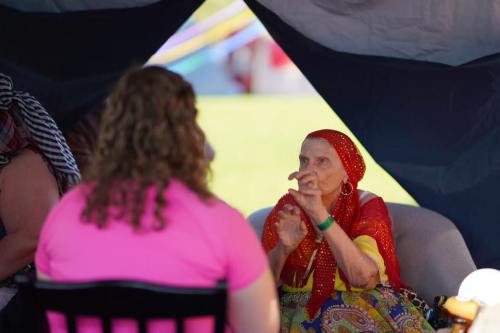 A woman in a colorful headscarf gestures while speaking to another person in a tent setting.
