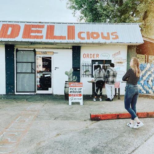A small deli with a sign reading "DELIcious," featuring customers ordering at a window and a woman standing nearby.