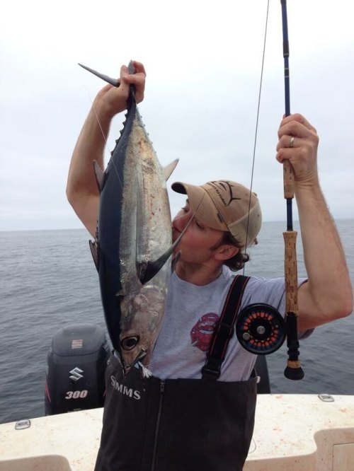 A fisherman on a boat holds a large fish, kissing it while holding a fishing rod in the other hand.