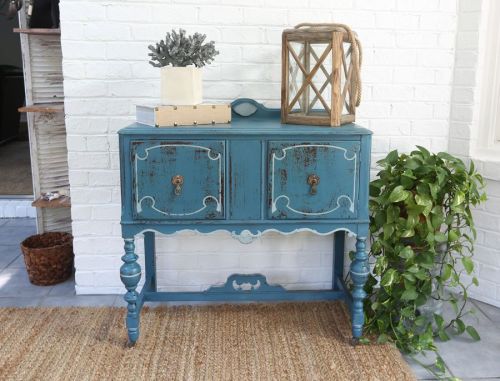 A vintage blue side table with two drawers, a lantern, and a plant, set against a white brick wall.