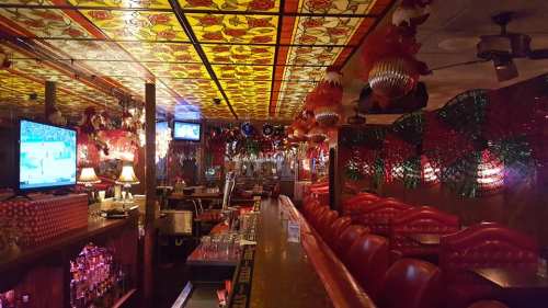 A vibrant bar interior with colorful stained glass ceiling, festive decorations, and empty red booths.