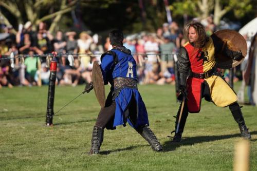 Two armored knights engage in a sword fight at a medieval event, with a crowd watching in the background.