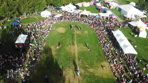 Aerial view of a large crowd at a Renaissance fair, with tents and a grassy area for activities.