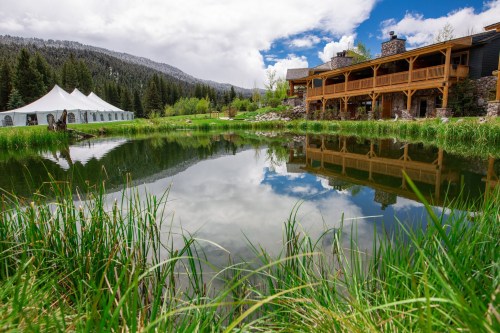 A serene pond reflects a cloudy sky, surrounded by lush grass and a rustic building with a tent nearby.