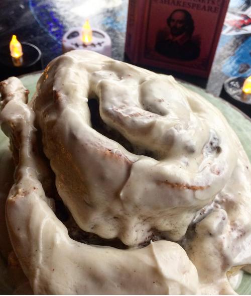 A large, frosted cinnamon roll sits on a plate, with candles and a book by Shakespeare in the background.