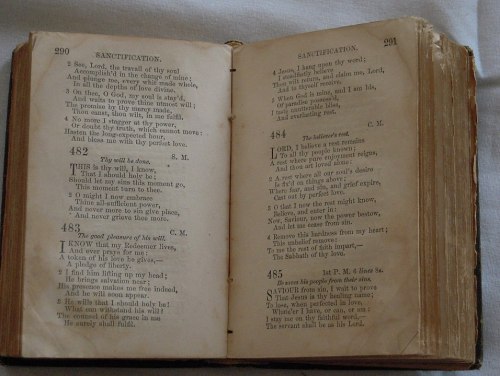 Open vintage hymn book pages with text, including titles and numbers for hymns, showing signs of wear and age.