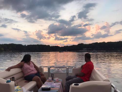 Two people sit on a boat, gazing at a colorful sunset over a calm lake, surrounded by trees and clouds.