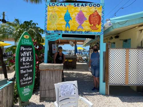 A colorful seafood restaurant entrance with a sign, two people waiting, and outdoor seating under yellow umbrellas.