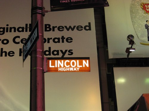 A street sign for Lincoln Highway illuminated against a large advertisement in Times Square.