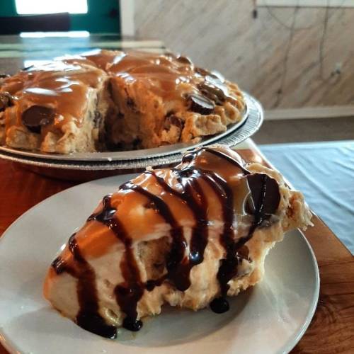 A slice of dessert topped with chocolate and caramel sauce, next to a whole pie on a table.