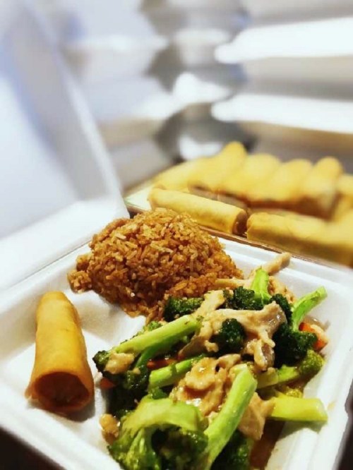 A takeout container with fried rice, chicken and broccoli, and a spring roll, with additional spring rolls in the background.