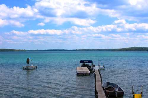 A serene lake scene with boats docked, a wooden pier, and fluffy clouds reflecting in the turquoise water.