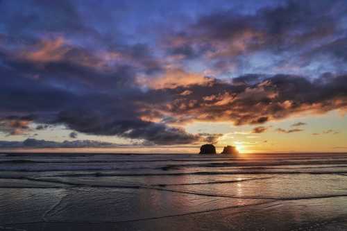 A serene beach at sunset, with colorful clouds reflecting on the water and two rock formations in the distance.