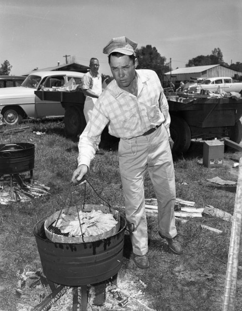 A man in a checkered shirt and cap cooks over a grill outdoors, with another person and vintage cars in the background.