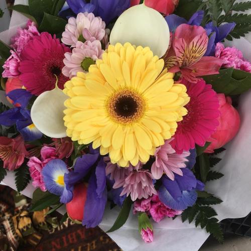 A vibrant bouquet featuring yellow gerbera daisies, pink and purple flowers, and lush green foliage.