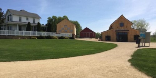 A scenic farm with a white house, two wooden barns, and a gravel path surrounded by green grass and trees.