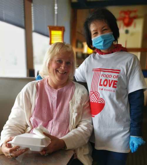Two women smiling in a restaurant, one wearing a "Food of Love" shirt and a mask, holding a takeout container.