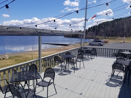 A lakeside deck with empty tables and chairs, string lights overhead, and a view of the water and distant hills.