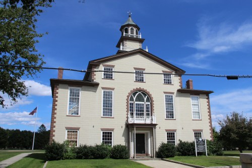 Historic building with a clock tower, surrounded by greenery and a blue sky with clouds. An American flag is visible.