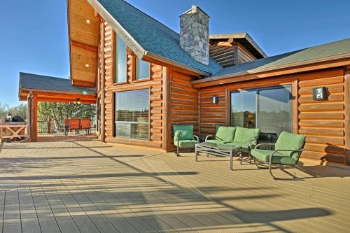 A log cabin exterior with a spacious deck, green chairs, and a stone chimney under a clear blue sky.