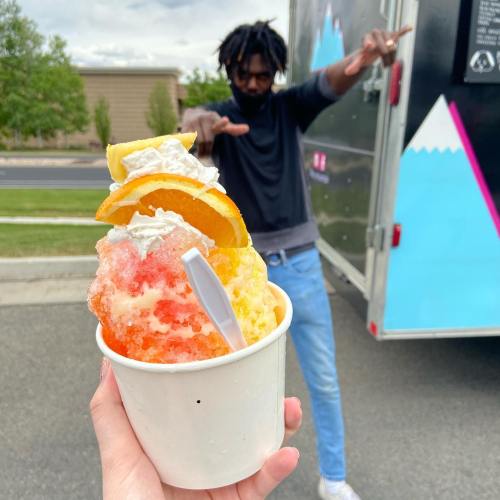A hand holds a colorful snow cone topped with fruit and whipped cream, with a person playfully posing in the background.