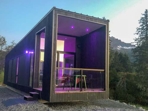 A modern black cabin with purple lighting, featuring a balcony and surrounded by trees and mountains.