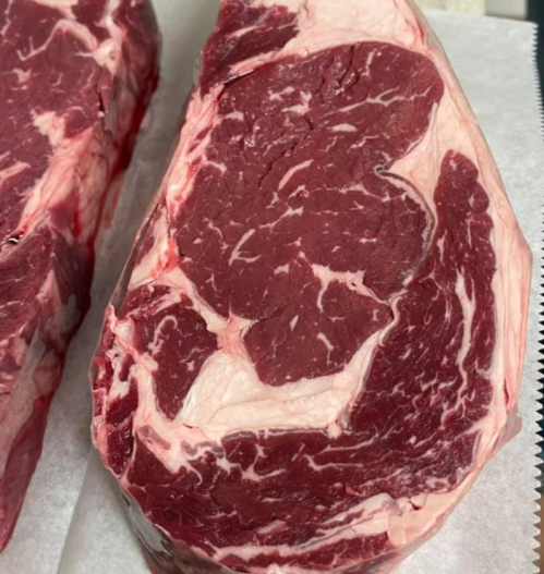 Close-up of two marbled cuts of raw ribeye steak on a white surface.