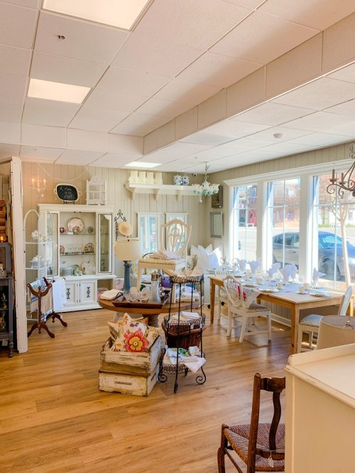 Bright, cozy interior of a shop featuring wooden furniture, decorative items, and large windows letting in natural light.