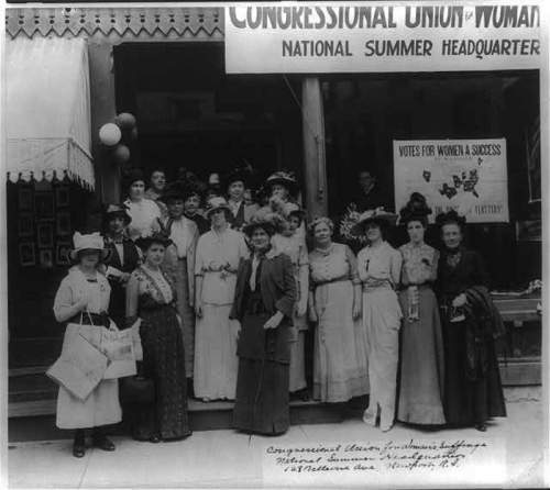 A historic group of women in early 20th-century attire, gathered outside a building for women's suffrage.