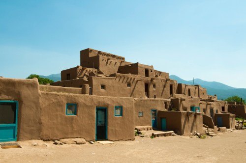 Adobe-style buildings with blue doors, set against a clear sky and mountains in the background.
