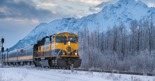 A yellow and blue train travels through a snowy landscape with mountains in the background and trees covered in frost.