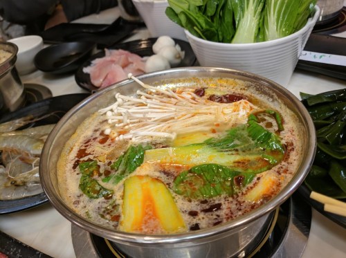 A steaming pot of spicy soup with vegetables, mushrooms, and seafood, surrounded by fresh ingredients on a table.