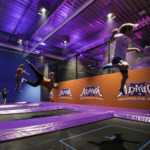 Two children jumping on trampolines in a brightly lit indoor trampoline park with purple lighting.