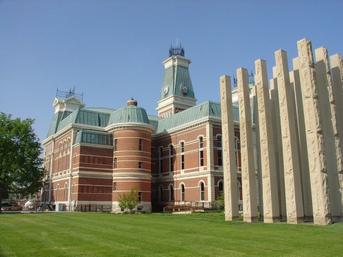 Historic brick building with a green copper roof, clock tower, and modern stone structure beside it, set against a clear blue sky.