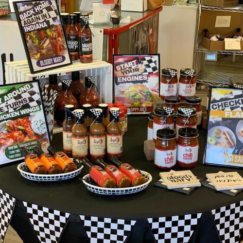 A display of sauces and snacks, including Bloody Mary mix, chicken wing sauce, and sloppy joe mix, on a checkered tablecloth.