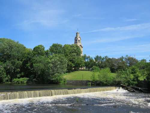 A serene landscape featuring a waterfall in the foreground and a historic building surrounded by lush greenery in the background.