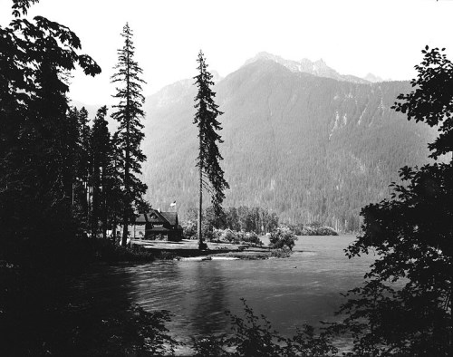 Black and white landscape featuring a cabin by a lake, surrounded by tall trees and mountains in the background.