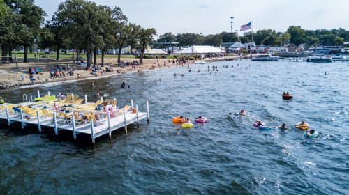 A busy beach scene with people swimming and relaxing on a dock, surrounded by trees and a festive atmosphere.