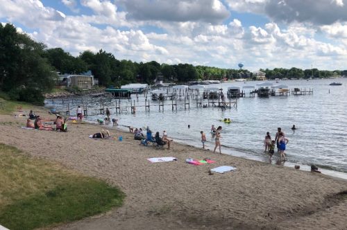 A sandy beach with people swimming and relaxing, boats docked nearby, under a partly cloudy sky.