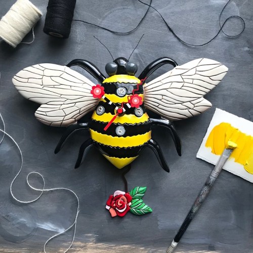 A decorative bee clock with yellow and black stripes, red flowers, and a rose, surrounded by thread and paint.