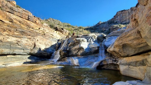 A serene rocky landscape with a small waterfall cascading into a clear pool, surrounded by desert vegetation and blue sky.