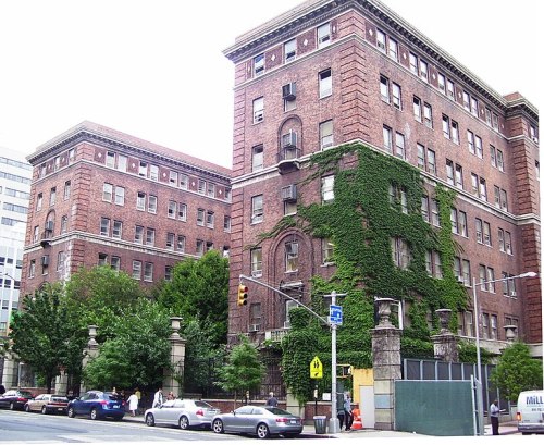 Historic brick building with ivy, featuring multiple stories and large windows, surrounded by trees and city streets.
