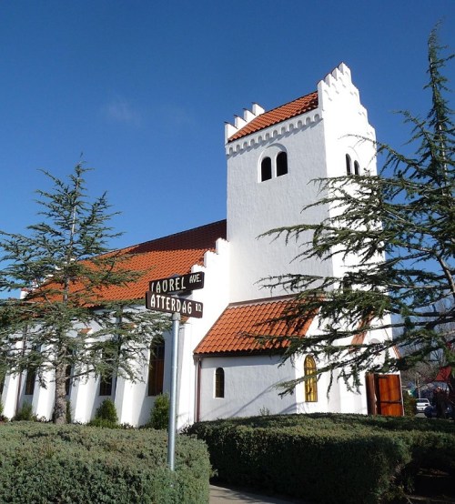 A white church with a red-tiled roof, surrounded by green shrubs and trees, under a clear blue sky.