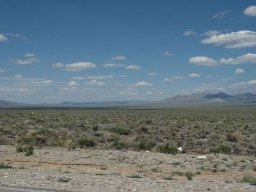 A vast, open landscape with sparse vegetation, mountains in the distance, and a blue sky dotted with clouds.