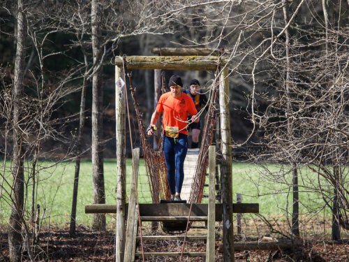 A runner in an orange shirt crosses a wooden suspension bridge surrounded by trees and a grassy field.