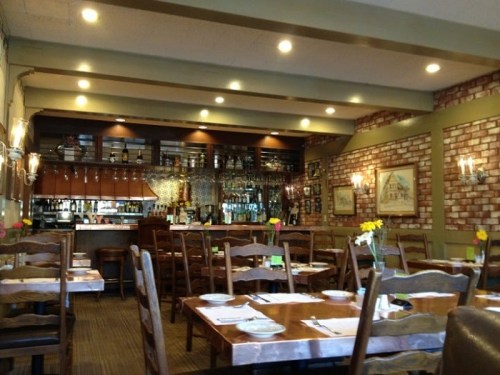 Cozy restaurant interior with wooden tables, chairs, and a bar area, featuring warm lighting and floral centerpieces.