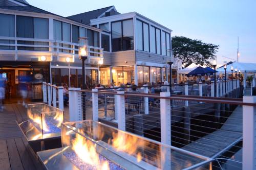 A waterfront restaurant with outdoor seating, illuminated by flames in glass containers, under a twilight sky.