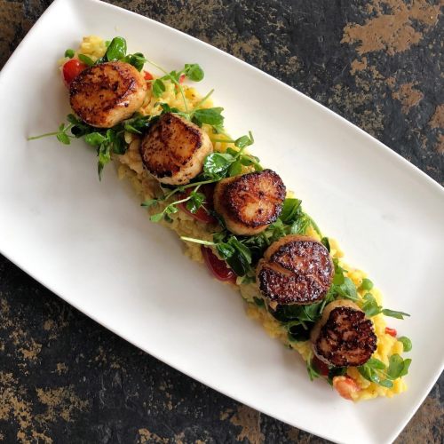 A long white plate with five seared scallops atop a bed of risotto, garnished with arugula and cherry tomatoes.